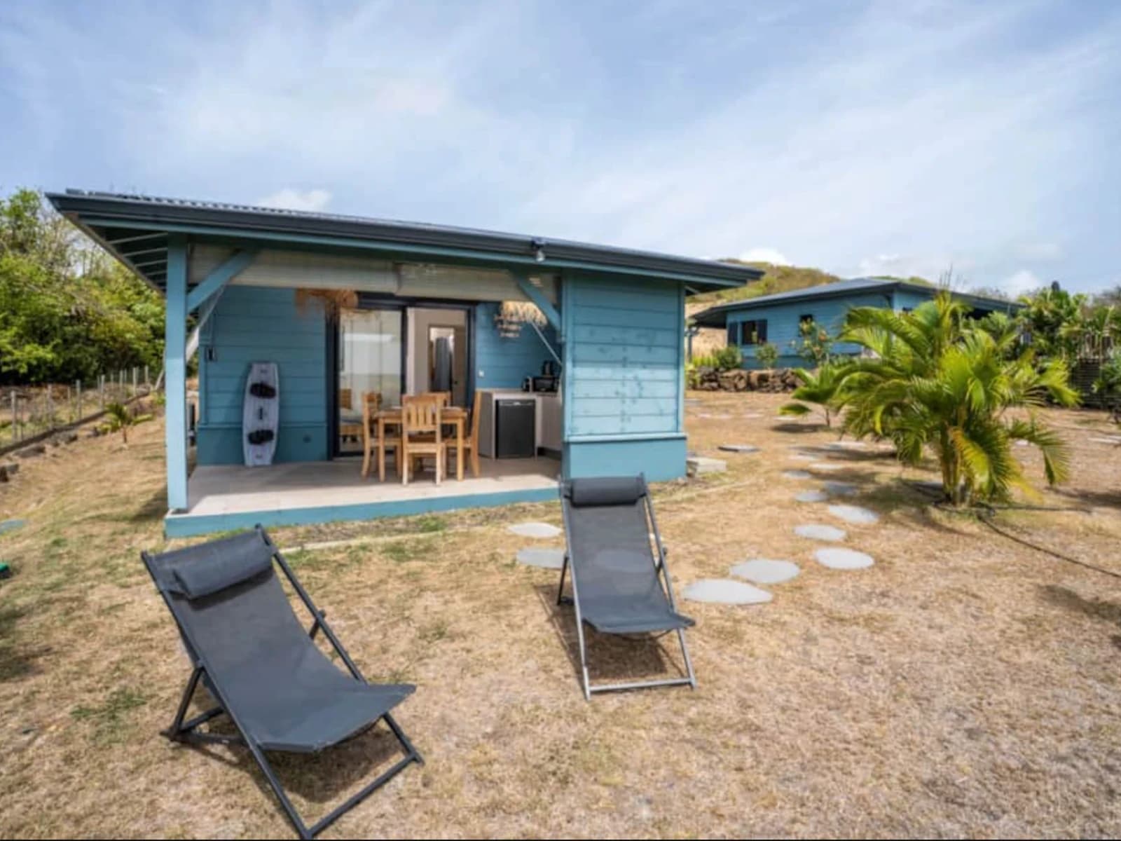 Bungalow en bois bleu avec terrasse couverte, deux transats sur la pelouse et palmiers